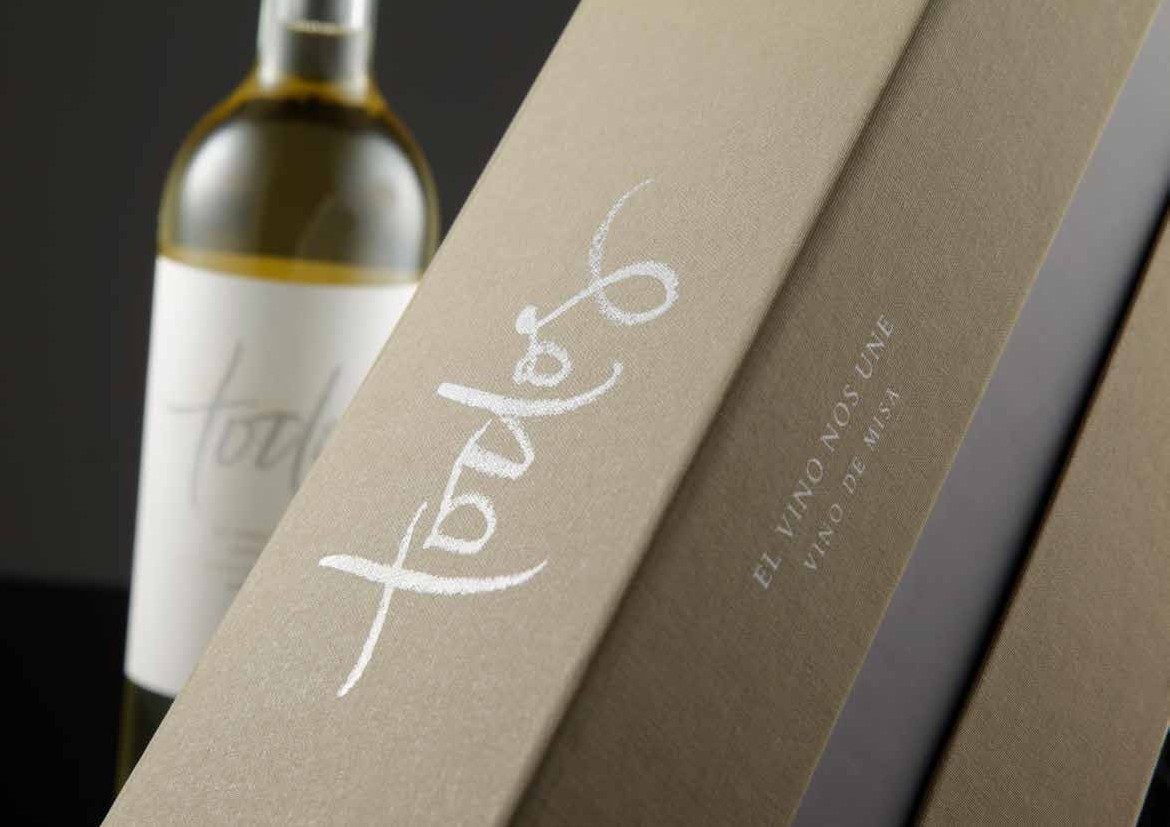 Estuche rígido forrado en papel entelado, con caligrafía blanca y texto en relieve, junto a una botella de vino